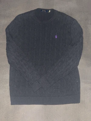 Mörkgrå stickad tröja Polo Ralph Lauren - Snygg mörkgrå stickad tröja från Polo Ralph Lauren med klassiskt kabelstickat mönster och lila broderad logga på bröstet. Tröjan har rund halsringning och ribbade muddar vid ärmslut och nederkant. Perfekt för lager på lager under kalla dagar.