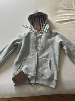 Grå zip hoodie från Burberry - Snygg grå zip hoodie från Burberry med klassiskt rutigt foder i huvan. Hoodien har dragsko i huvan, två stora fickor fram och ribbade muddar. Perfekt för dig som gillar exklusiva streetwear-plagg och vill ha något som sticker ut. Säljer den för att den var för liten.