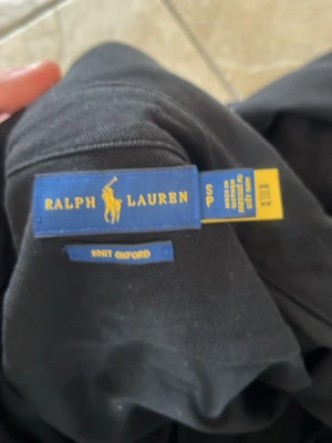 Svart skjorta från Ralph Lauren - Klassisk svart skjorta från Ralph Lauren med knappkrage och broderad logga på bröstet. Tillverkad i mjuk bomull med långa ärmar och vita knappar. Modellen är en Knit Oxford som ger en avslappnad men ändå stilren look.