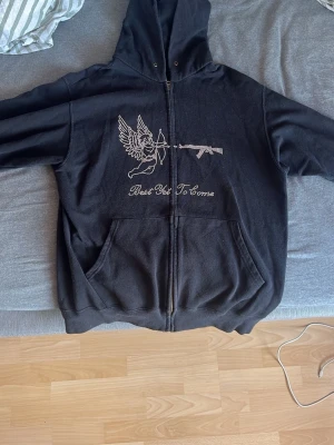Rhinestone Hoodie, best yet to come - Svart hoodie med dragkedja och huva. På framsidan finns ett stort rhinestone tryck med en bevingad ängel som skjuter pilbåge mot ett gevär och texten 'Best Yet To Come'. Perfekt för dig som gillar streetwear och unika detaljer. Storlek är M men är liten i storlek