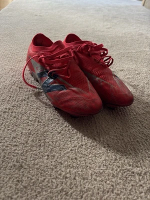 Röda New Balance fotbollsskor - Säljer ett par röda fotbollsskor från New Balance med snörning och låg profil. Skorna har en stor silverfärgad och svart logga på sidan och är tillverkade i ett lätt syntetmaterial som ger bra bollkontroll. Perfekta för dig som vill ha speed och stil på planen.