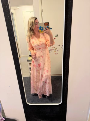 Ljusrosa långklänning med blommor - Romantisk långklänning i ljusrosa med blommigt mönster och v-ringning. Klänningen markerad midja, vilket ger en feminin siluett. Perfekt för vår och sommar. Materialet känns lätt och luftigt.Storlek 38/40 märkt som M.Helt ny.