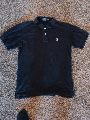 Svart piké från Polo Ralph Lauren - Klassisk svart pikétröja från Polo Ralph Lauren med vit broderad logga på bröstet. Tröjan har korta ärmar, krage och två knappar framtill. Tillverkad i mjuk bomull och har en normal passform. Perfekt till en avslappnad men ändå stilren look.