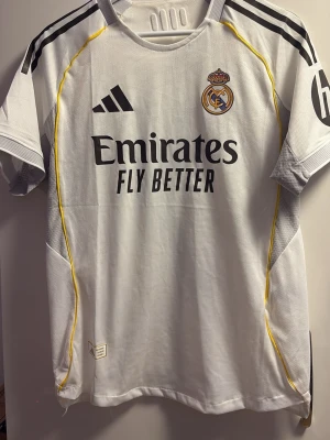 Real Madrid matchtröja Vini Jr. 7 - Säljer en vit Real Madrid matchtröja från Adidas med Vini Jr. och nummer 7 på ryggen. Tröjan har svarta detaljer, gula linjer längs sidorna och klubbmärke på bröstet. Tillverkad i lätt och ventilerande material, perfekt för fotbollsträning eller match.