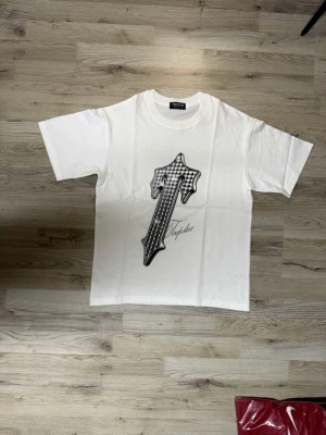 Trapstar T shirt - Säljer en vit T-shirt från Trapstar med ett stort svartvitt T-tryck framtill i rutigt mönster och stilren text under. Perfekt för en clean och trendig look.