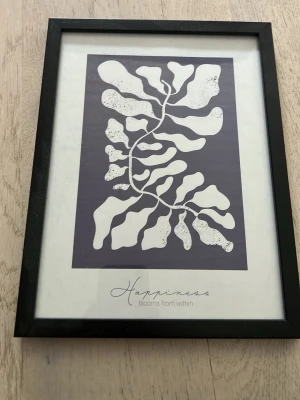 Tavla med svart ram och växtmotiv - Snygg tavla med svart ram och abstrakt växtmotiv i vitt mot mörklila bakgrund. Under motivet står texten 'Happiness blooms from within'. Perfekt som dekoration till rummet eller kontoret. Ramen är av plast och har stöd på baksidan.