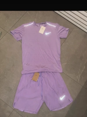 Lila träningsset från Nike - Säljer ett lila träningsset från Nike med t-shirt och shorts. Båda plaggen har reflekterande Nike-logga och är tillverkade i ett lätt och snabbtorkande material, perfekt för löpning eller gym. T-shirten har rund hals och shortsen har dragsko i midjan.