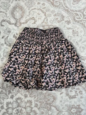 Blommig kortkjol från Gina Tricot - Supersöt kortkjol från Gina Tricot med blommigt mönster i svart, rosa och beige. Kjolen har bred smock i midjan för en bekväm passform och ett flowy fall. Perfekt för att addera lite färg till din outfit!                        Den är insydd i midjan men går att sprätta upp till den orginala storleken! 