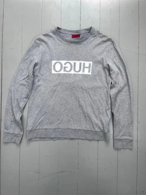 Grå sweatshirt från Hugo Boss - Snygg grå sweatshirt från Hugo Boss med vit HUGO-logga tryckt på bröstet. Tröjan har rund halsringning, långa ärmar och ribbade muddar. Perfekt för en avslappnad och trendig stil.
