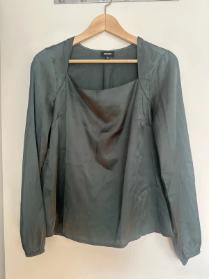 Mörkgrön satinskjorta från BUBBLEROOM - Stilren mörkgrön blus från BUBBLEROOM i glansigt satintyg. Blusen har långa ärmar med lätt puff och en snygg draperad fyrkantig ringning. Perfekt för dig som gillar en enkel men trendig look. Den har några lösa tråd men märks vid nära inspektion, även en liten fläck som visas i bilderna.