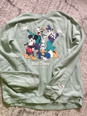 Grön Disney sweatshirt med tryck - Snygg ljusgrön sweatshirt från Disney med stort tryck av Musse Pigg, Kalle Anka, Långben och Kajsa Anka på framsidan. Rund halsringning, ribbade muddar och mjukt material. Texten 'Walt Disney EST 1928' under motivet. Perfekt för dig som älskar klassiska Disney-karaktärer.