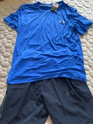 Blå träningsset från Under Armour - Säljer ett sportigt träningsset från Under Armour med en blå t-shirt och svarta shorts. T-shirten har rund hals och logga på bröstet, shortsen har diskret logga och sidofickor. Perfekt för gym eller löpning. Materialet är lätt och andas.