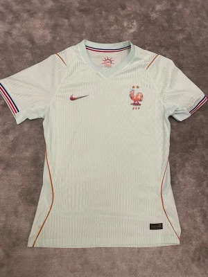 Frankrike Nike fotbollströja ljusblå - Snygg ljusblå fotbollströja från Nike med Frankrikes landslagsemblem och två stjärnor på bröstet. Tröjan har korta ärmar med rödblå-vita detaljer och orangea linjer längs sidorna. Perfekt för fotbollsträning eller match.