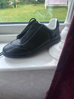 Maison Margiela Future - Svarta sneakers från Maison Margiela med minimalistisk design och diskreta sömmar. Skon har en låg siluett, svarta skosnören och vit insida. Ovandelen är i slätt läder och yttersulan är i gummi med tydlig logotyp under sulan. Om det finns några frågor så skriv tack!