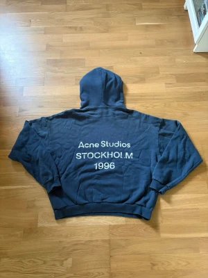 Mörkblå hoodie Acne Studios - Säljer en mörkblå hoodie från Acne Studios med stor huva och tryck på ryggen: 'Acne Studios STOCKHOLM 1996'. Hoodien har en avslappnad passform och är gjord i mjuk bomull. Perfekt för dig som gillar streetwear och vill ha något stilrent men ändå unikt. Acne plastpåsen medföljer ej på denna.