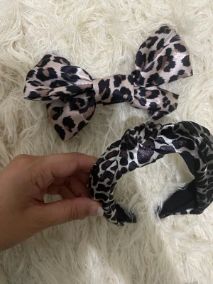 Leopardmönstrat hårband & rosett - Säljer ett set med ett brett hårband och en stor rosett med hårklämma, båda i glansigt tyg med leopardmönster i svart och beige. Perfekta accessoarer för att ge din look en trendig touch. Materialet känns mjukt och följsamt.