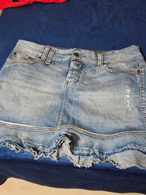 Blå jeanskjol med fransig kant Object - Säljer en blå jeanskjol från Object med fransig, rå kant och slitna detaljer. Kjolen har klassiska fem fickor, bälteshällor och knappgylf. Perfekt för dig som gillar en avslappnad och trendig stil.