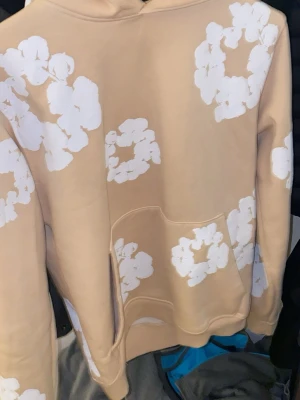 Beige hoodie med vita blommor Denim Tears - Säljer en beige hoodie från Denim Tears med stora vita blommönster över hela plagget. Hoodien har en klassisk huva och en stor magficka framtill. Perfekt för dig som vill sticka ut med en unik och trendig design.