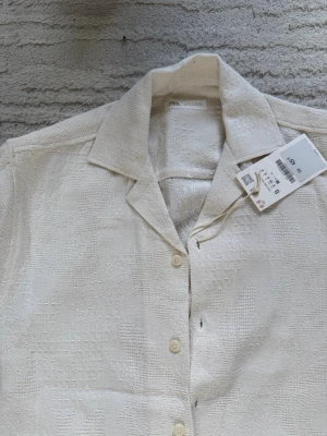 Beige kortärmad skjorta från Zara - Stilren beige skjorta från Zara i kortärmad modell. Skjortan har ett diskret strukturerat mönster, klassisk krage och knäppning framtill. Perfekt för en avslappnad men snygg look. Materialet känns lätt och luftigt.