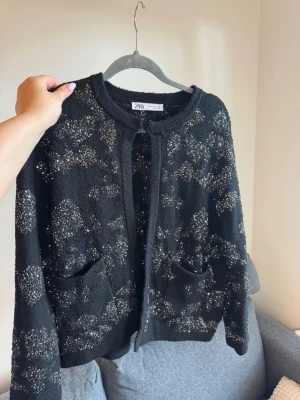Svart glittrig kofta från ZARA - Snygg svart kofta från ZARA med glittriga silverdetaljer i ett mönster över hela plagget. Koftan har rund halsringning, två framfickor och är öppen framtill utan knappar. Det är storlek Xl men är mer som en s-m