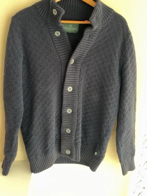 Marinblå stickad cardigan från Kronstadt - Marinblå stickad cardigan med ribbad krage och knappslå. Koftan har ett diskret rutmönster, långa ärmar och breda ribbade muddar vid ärmslut och nederkant. Perfekt för lager på lager och en stilren look. Perfekt för sommarkvällar. Hör av er vid funderingar!
