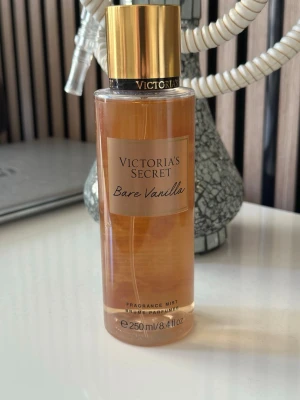 Victoria's Secret Bare Vanilla mist - En lyxig doftmist från Victoria's Secret i doften Bare Vanilla. Flaskan är cylindrisk och genomskinlig med guldigt lock och beige etikett. Innehåller 250 ml och har en varm, söt vaniljdoft som känns både mjuk och modern.