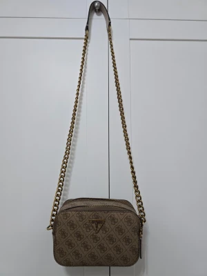 Brun axelväska från Guess med kedja - Snygg brun axelväska från Guess med guldfärgad kedjerem och klassiskt logomönster över hela väskan. Väskan har en rektangulär form, dragkedja upptill och Guess metallogo framtill. 