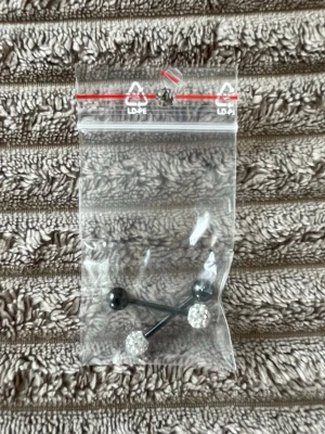 Svarta tungpiercing med glittrande pärlor - Säljer en tungpercing med svart stav och en kula i svart samt en kula täckt av glittrande vita pärlor. Perfekta för dig som gillar en mix av klassiskt och trendigt. Kommer i en liten plastpåse.