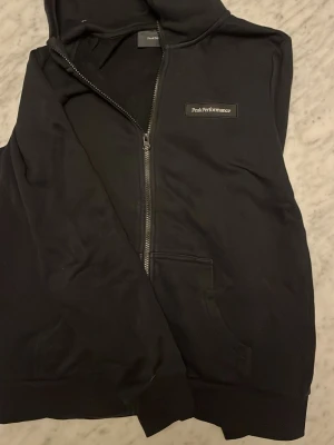 Svart hoodie från Peak Performance - Säljer en svart hoodie från Peak Performance med dragkedja framtill och fickor på sidorna. Hoodien har en liten logga på bröstet och är tillverkad i mjukt bomullsmaterial. Perfekt för en avslappnad och stilren look. Den är för liten för mig vilket syns men den är M 170/92A