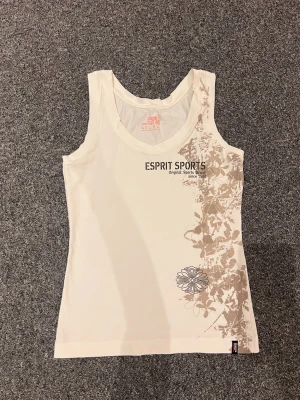 Vit linnetopp från Esprit Sports - Snygg vit linnetopp från Esprit Sports med beige blommigt mönster på ena sidan och tryck framtill. Perfekt för en sportig och avslappnad stil. Mjuk och skön bomullskvalitet med ärmlös design och rund halsringning.