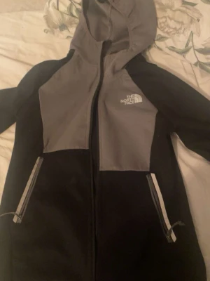 The North Face kofta  - The North Face huvjacka i svart och grått med huva, dragkedja och två fickor med dragkedja. Sportig och bekväm modell som passar både vardag och fritid.
