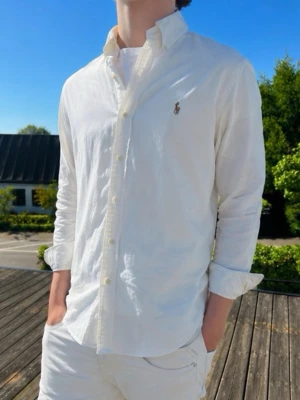 Vit skjorta från Ralph Lauren - Klassisk vit skjorta från Ralph Lauren med button-down krage och broderad logga på bröstet. Skjortan har lång ärm, knappar framtill och är tillverkad i mjuk bomull. Perfekt för en clean och stilren look. Bvsa!