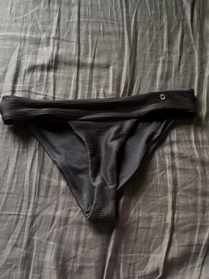 Svarta ribbade bikinitrosor - Snygga svarta bikinitrosor med ribbad struktur och en liten metallogo framtill. Klassisk modell med bredare kant upptill som ger en stilren look. Perfekt för strand eller poolhäng.