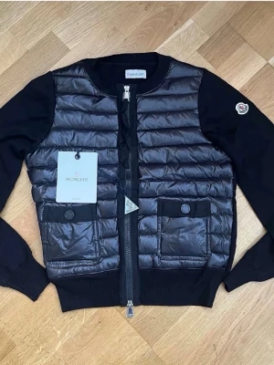 Svart cardiganjacka från Moncler - Snygg svart cardiganjacka från Moncler med quiltad, glansig front och stickade ärmar. Jackan har två stora fickor framtill med knappstängning och dragkedja i mitten. Moncler-logga på ärmen och ribbade muddar för extra komfort. Kvitto och tags medföljer.
