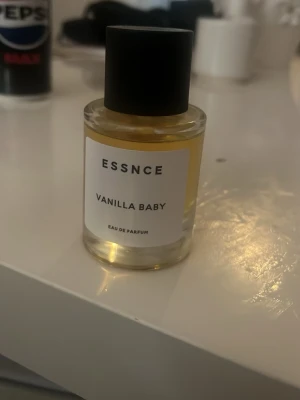 ESSNCE Vanilla Baby Eau de Parfum - En stilren parfymflaska från ESSNCE med doften Vanilla Baby. Flaskan är cylindrisk i genomskinligt glas med svart lock och enkel vit etikett. Innehållet är ljusgult och ger en fräsch och modern känsla, perfekt för dig som gillar söta vaniljtoner.