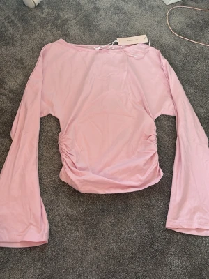 Ljusrosa långärmad topp med rynk - Säljer en ljusrosa långärmad topp med rynkade detaljer i sidorna. Toppen har en enkel, stilren design med rund halsringning och är tillverkad i ett mjukt material som känns skönt mot huden. Perfekt för dig som gillar en feminin och trendig look.