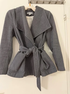 Grå kappa med knytskärp - Stilren grå kappa med bred krage och knytskärp i midjan. Kappan har två stora fickor framtill och är tillverkad i ett mjukt, ull-liknande material. Perfekt för kyliga dagar och ger en snygg siluett.