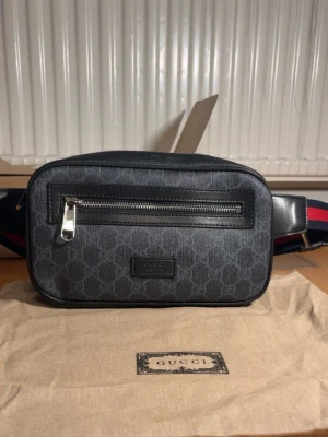 GG axelväska med monogram - Snygg axelväska från GG i mörkgrått med klassiskt GG-monogram. Väskan har en framficka med dragkedja, svarta läderdetaljer och justerbar axelrem i blått och rött. Materialet är canvas med läderdetaljer och baksidan har mesh för extra komfort.