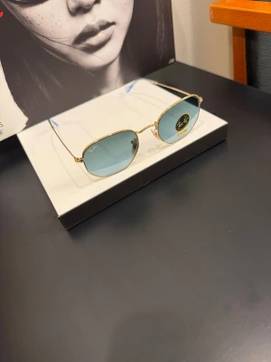 Ray-Ban solglasögon guld blå - Snygga Ray-Ban solglasögon med tunna guldfärgade metallbågar och blåtonade glas. Modellen har en rektangulär form som ger en modern och stilren look. Perfekta för dig som vill ha en klassisk accessoar med en twist.