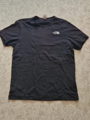 Svart t-shirt från The North Face - Svart t-shirt från The North Face med liten vit logga på bröstet och stor röd/vit logga samt texten 'Never Stop Exploring' på ryggen. Klassisk passform och rund halsringning. Perfekt till vardags för en clean och sportig look.