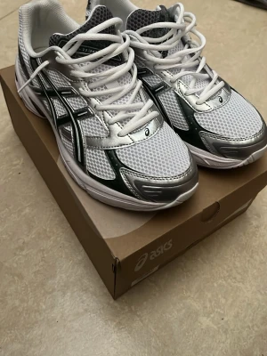 Asics gel 1130 - Snygga Asics sneakers i vitt meshmaterial med detaljer i silver och mörkgrönt. Klassisk låg modell med vita skosnören och svart Asics-logga på sidan. Perfekta för dig som gillar sportig och trendig stil.