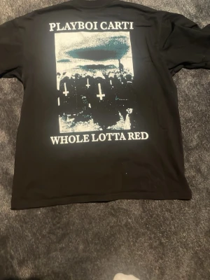 Playboi Carti Whole Lotta Red T-shirt - Svart t-shirt med tryck från Playboi Cartis album 'Whole Lotta Red'. Stora grafiska motiv och text på både fram- och baksida. Materialet känns som bomull och passformen är normal. Perfekt för dig som gillar streetwear och vill sticka ut. Storlek M men passar som S
