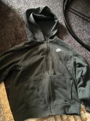Svart zip-hoodie från Nike - Säljer en svart hoodie från Nike med dragkedja framtill och två fickor. Klassisk Nike-logga broderad i vitt på bröstet. Hoodien har ribbade muddar och är tillverkad i mjukt bomullsmaterial. Perfekt för en avslappnad och sportig stil.