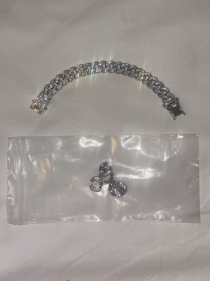 Silverfärgat armband med stenar - Kommer med extra om man vill göra de större. Säljer ett chunky armband i silverfärg med glittrande stenar på varje länk. Armbandet har ett brett kedjemönster och ett lås dekorerat med små stenar. Perfekt för dig som gillar statement-smycken och vill ha något som verkligen syns.
