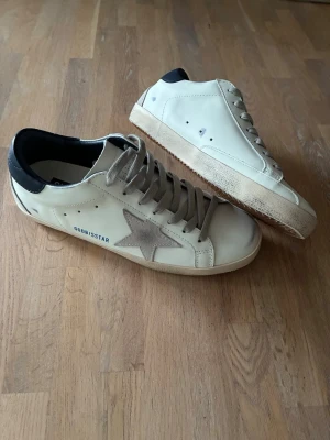 Golden Goose Superstar - Snygga vita sneakers från Golden Goose med grå mockadetaljer och den ikoniska stjärnan på sidan. Skorna har mörkblå hälkappa, grå skosnören och en vintage-inspirerad sula. Tillverkade i läder och mocka för en cool och avslappnad look.