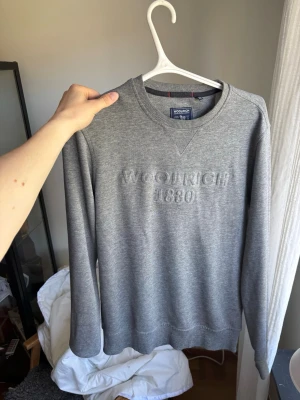 Grå sweatshirt från Woolrich - Snygg grå sweatshirt från Woolrich med diskret broderad logga och årtal '1830' på bröstet. Tröjan har rund halsringning, ribbade muddar och är tillverkad i mjukt bomullsmaterial. Perfekt för sena sommarkvällar!