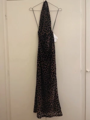 Leopardmönstrad långklänning - Snygg långklänning med leopardmönster i svart och beige. Klänningen har tunna axelband och en djup urringning framtill. Materialet känns mjukt och följsamt, troligen polyester eller liknande. Perfekt för dig som vill sticka ut med ett djuriskt print.
