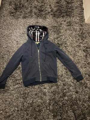 Marinblå hoodie med rutigt foder - Snygg mörkblå zip hoodie från Burberry. Huvan har ett klassiskt rutigt foder i svart, vitt och rött. Tillverkad i mjukt bomullsmaterial och har ribbade muddar vid ärmar och nederkant. Perfekt för en avslappnad och trendig look.