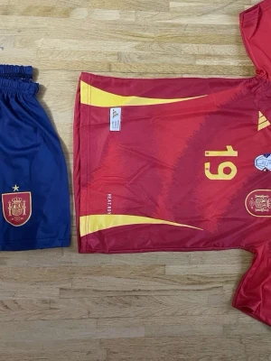 Spaniens matchställ Adidas nr 19 - Säljer ett matchställ från Spaniens landslag med röd tröja och gula detaljer, nummer 19 på både fram- och baksida samt blå shorts med landslagets emblem. Tillverkat i lätt och ventilerande material, perfekt för fotbollsträning eller match.