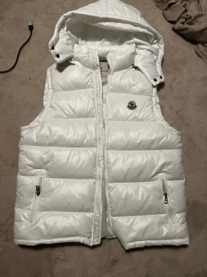 Vit dunväst med huva - Snygg vit dunväst med avtagbar huva och två fickor med dragkedja. Västen har en snygg logga på bröstet och quiltad design som ger en puffig look. Perfekt för lager-på-lager och håller dig varm under kyliga dagar.